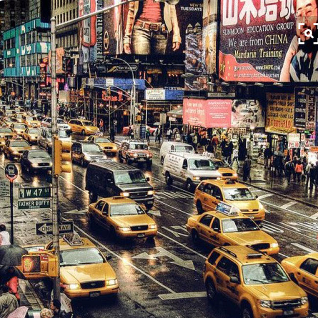 A busy city intersection with numerous yellow taxis, cars, and pedestrians, surrounded by tall buildings and colorful billboards.